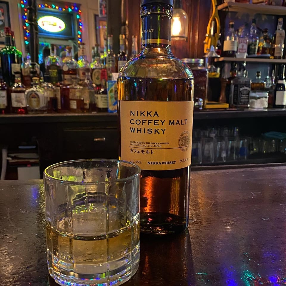 Nikka Coffey Malt Whisky bottle and tumbler on a colorful, warmly lit bar counter surrounded by liquor bottles.