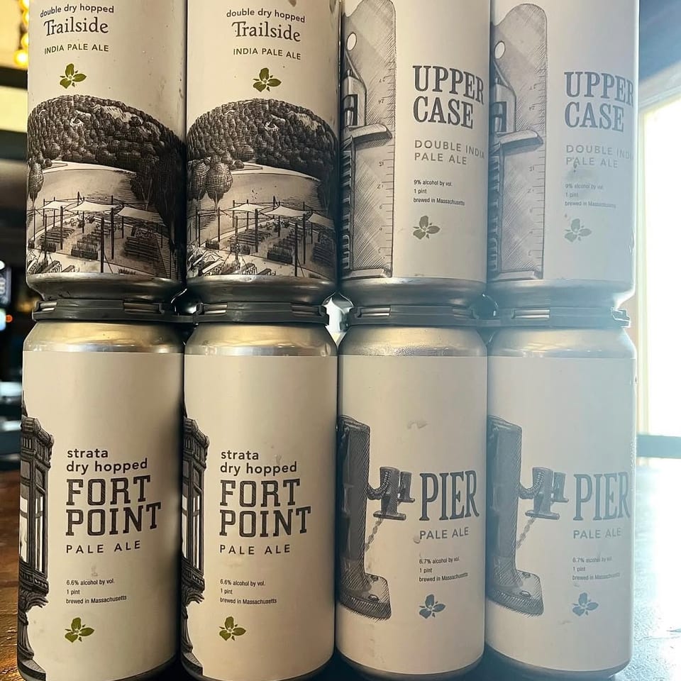 Stacked cans of Massachusetts-brewed pale ales, including Trailside, Upper Case, Fort Point, and Pier, displayed on a counter.
