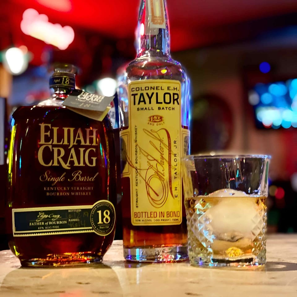Elijah Craig and Colonel E.H. Taylor bourbon bottles beside a whiskey cocktail in a dimly lit bar.